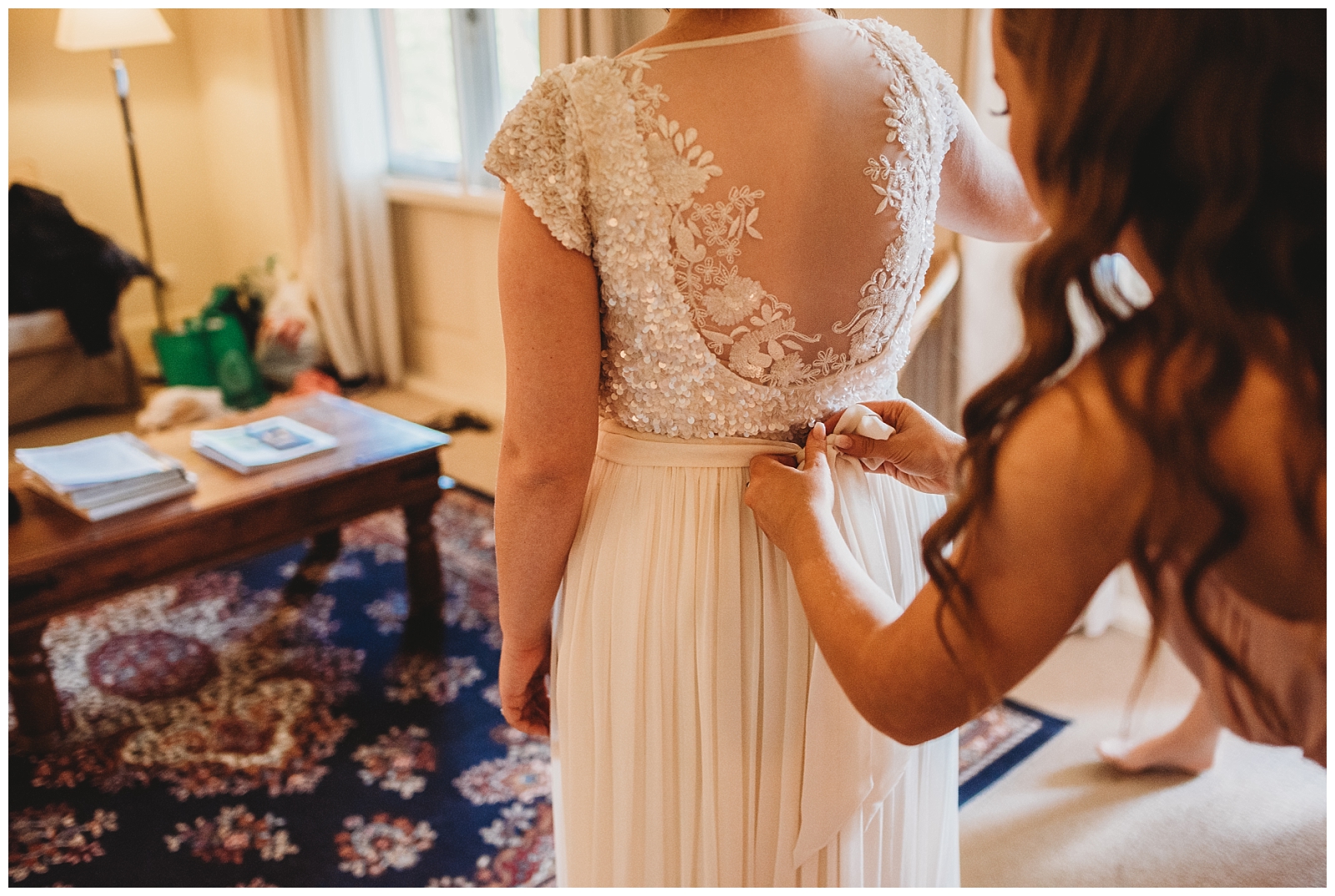 Bridesmaids hands tying ribbon on back of brides lace bodice dress.