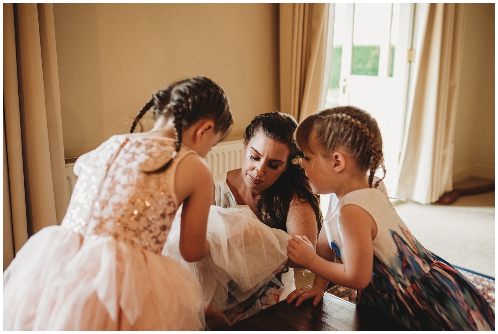 Woman and three children sit on floor as woman helps one child get flower girls dress on and the remaining two girls try to help.