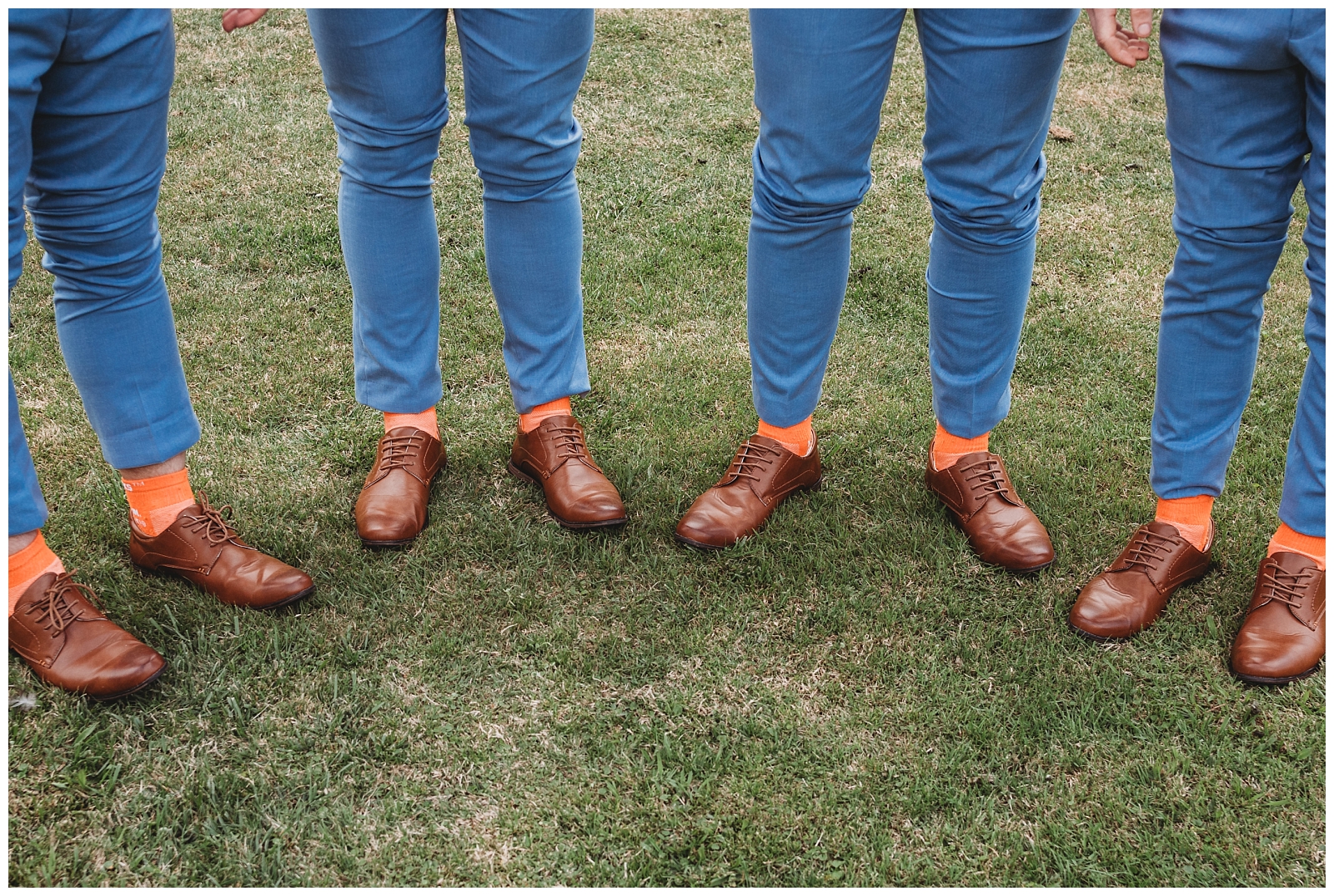 Groomsmen stand in arc shape lifting pant legs to show matching shoes and orange socks.