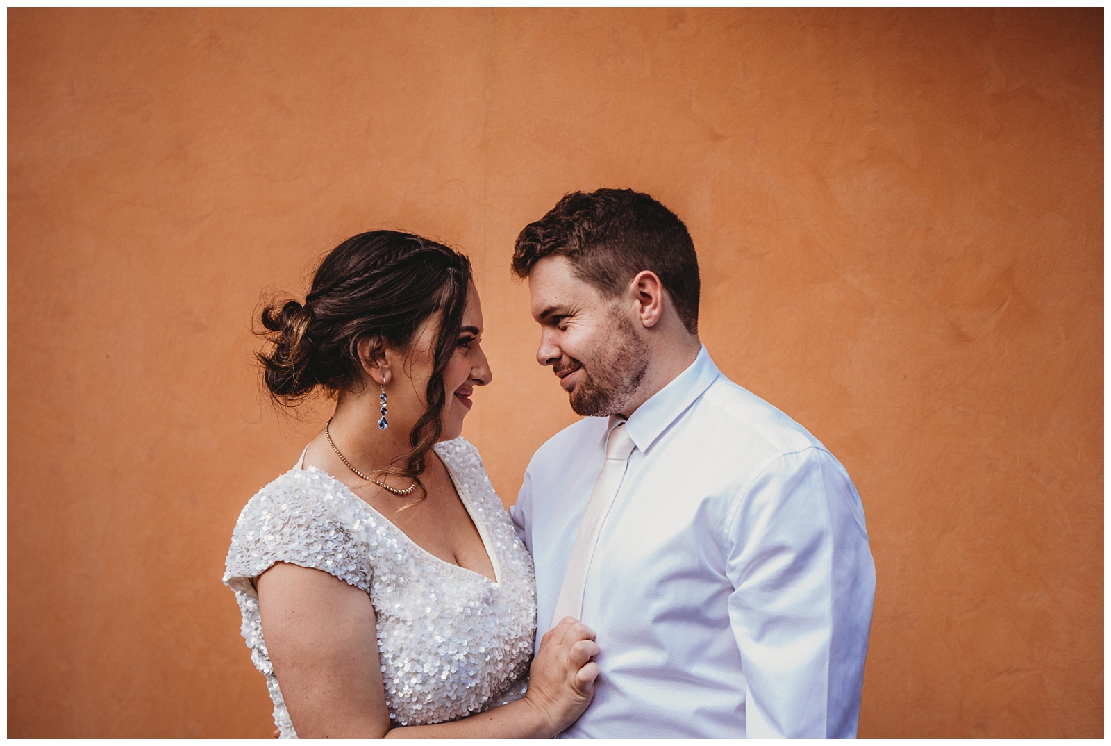 Brid and Groom pulled close together facing each other  in front of orange wall.
