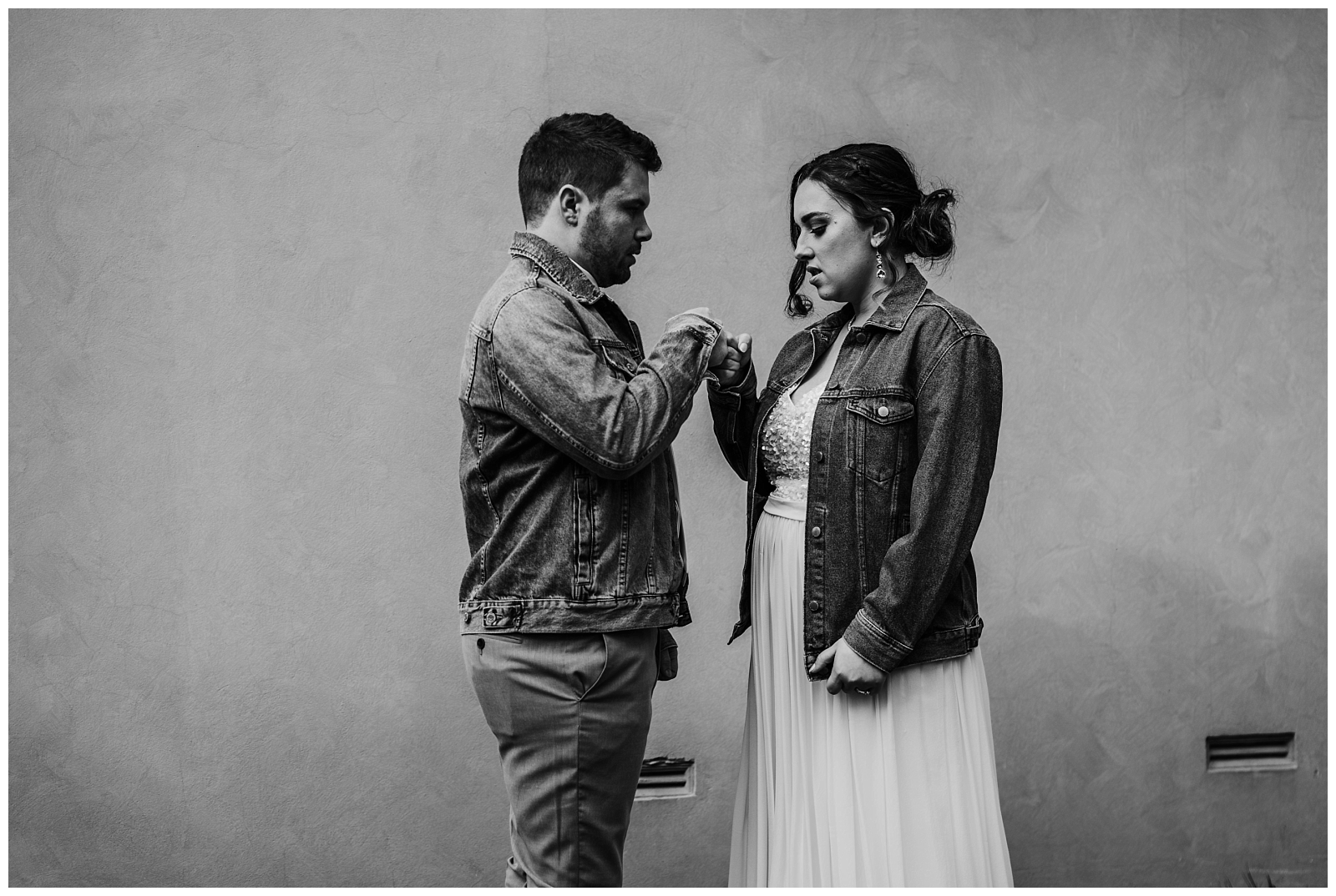 Bride and Groom in denim jackets stand facing each other doing fist bump in front of wall.