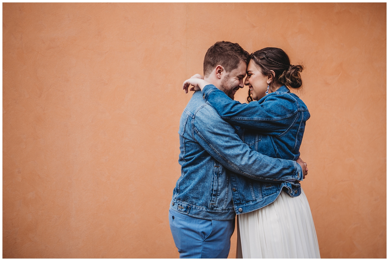 Bride and Groom wearing denim jackets stand in front of orange wall giggling with foreheads together.