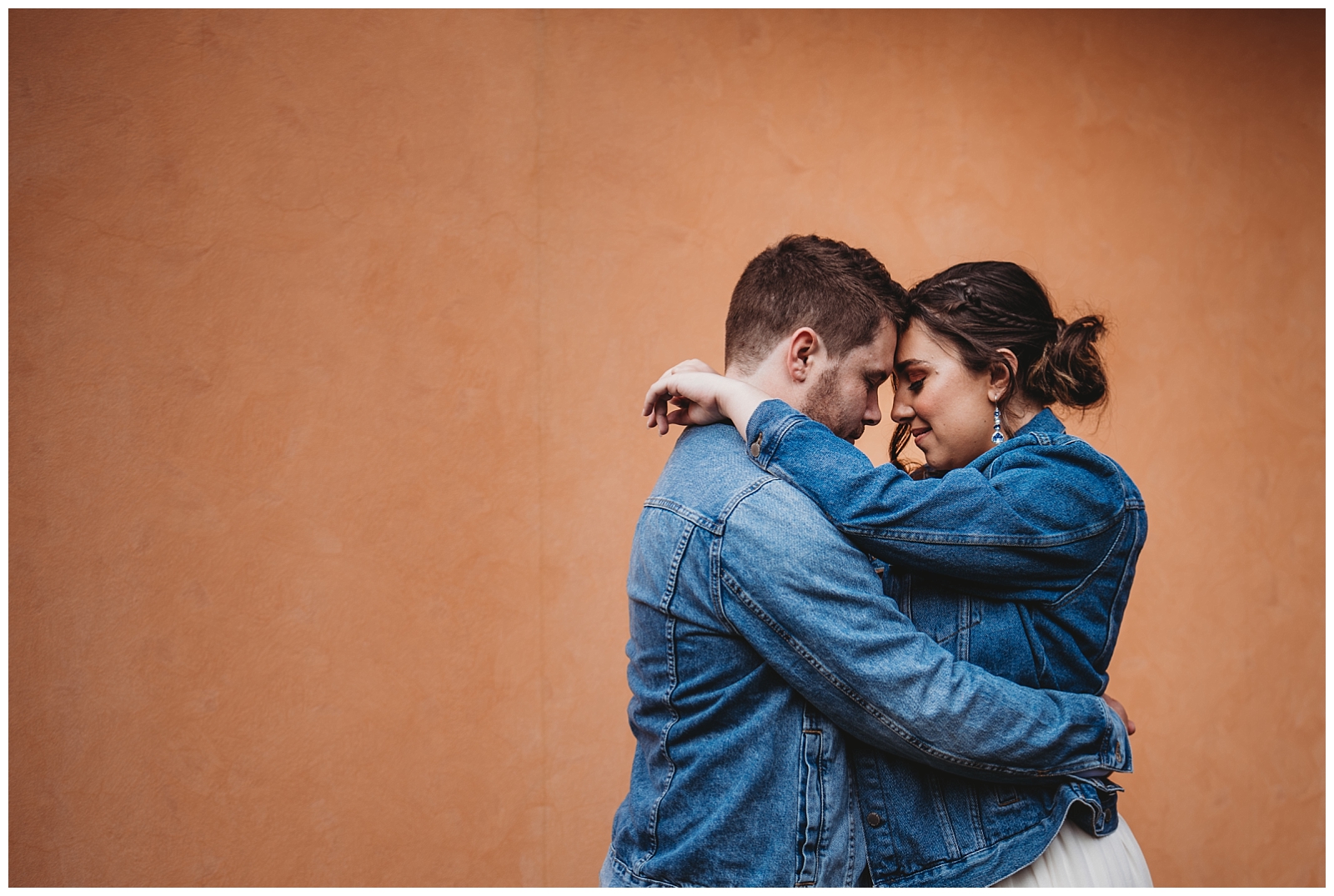 Bride and Groom in denim jackets stand facing each other with foreheads together in front of orange walll.
